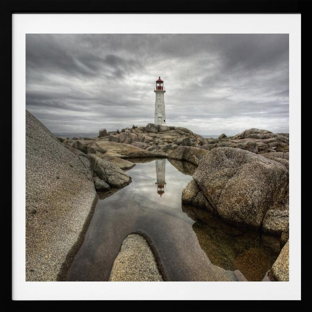 A framed photograph of a white lighthouse with a red top standing on a rocky shore under a dramatic, cloudy sky. In the foreground, a tide pool perfectly reflects the lighthouse. Artwork