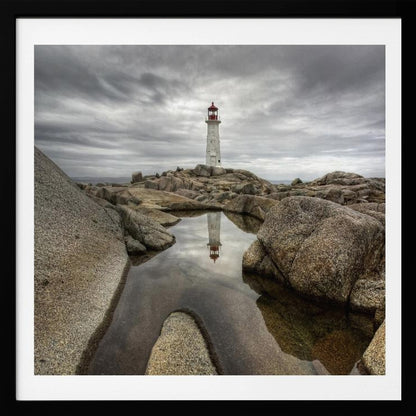 A framed photograph of a white lighthouse with a red top standing on a rocky shore under a dramatic, cloudy sky. In the foreground, a tide pool perfectly reflects the lighthouse. Artwork
