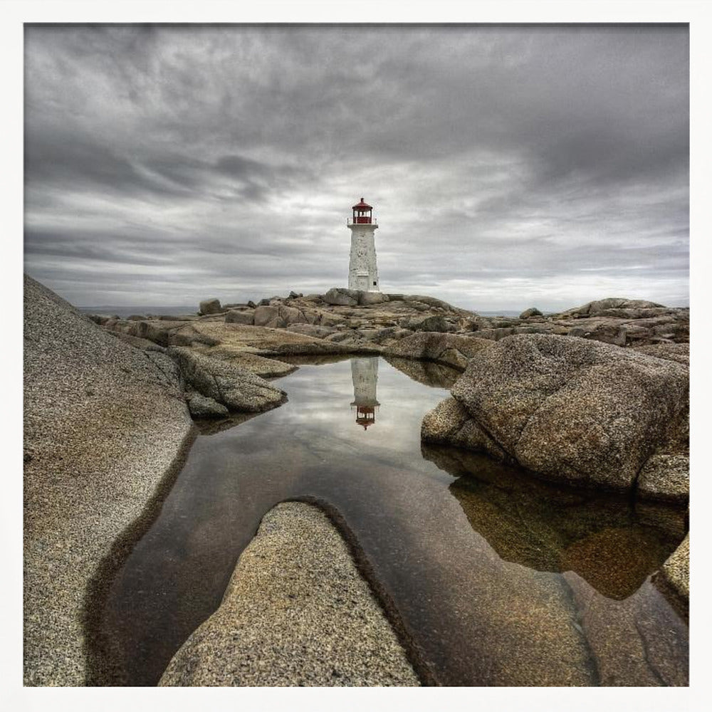A framed photograph of a white lighthouse with a red top standing on a rocky shore under a dramatic, cloudy sky. In the foreground, a tide pool perfectly reflects the lighthouse. Artwork