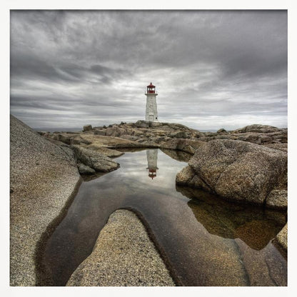 A framed photograph of a white lighthouse with a red top standing on a rocky shore under a dramatic, cloudy sky. In the foreground, a tide pool perfectly reflects the lighthouse. Artwork