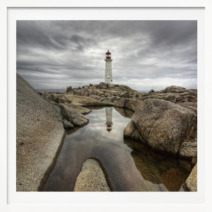 A framed photograph of a white lighthouse with a red top standing on a rocky shore under a dramatic, cloudy sky. In the foreground, a tide pool perfectly reflects the lighthouse. Artwork