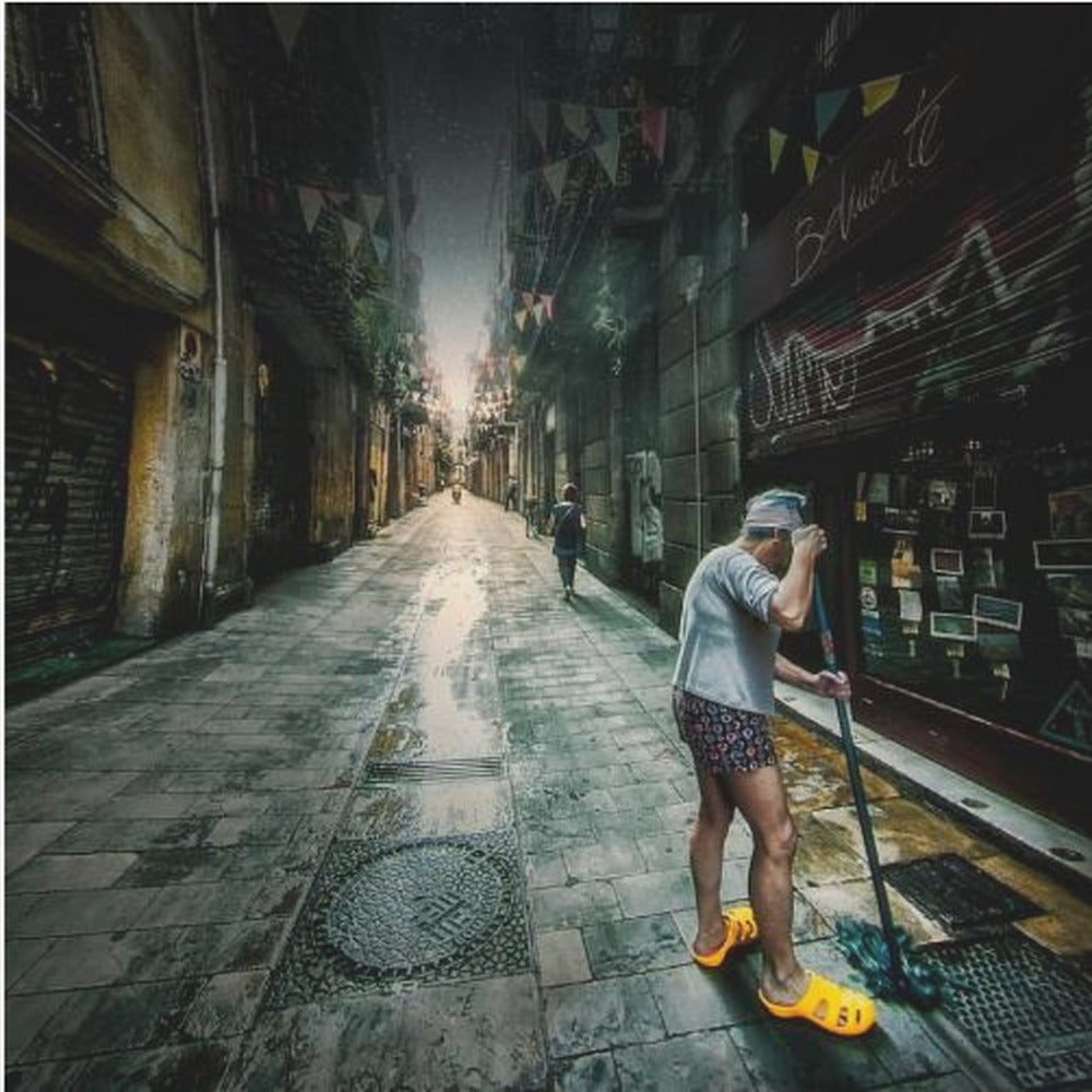 A wide-angle, atmospheric photograph of a man in bright yellow clogs mopping a wet, narrow cobblestone alley. The dark, gritty city street is lined with old buildings and closed storefronts, with another person walking in the distant light at the end of the alley. Wall Art