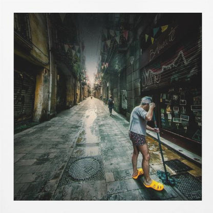 A wide-angle, atmospheric photograph of a man in bright yellow clogs mopping a wet, narrow cobblestone alley. The dark, gritty city street is lined with old buildings and closed storefronts, with another person walking in the distant light at the end of the alley. Wall Art