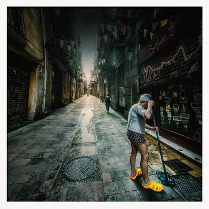 A wide-angle, atmospheric photograph of a man in bright yellow clogs mopping a wet, narrow cobblestone alley. The dark, gritty city street is lined with old buildings and closed storefronts, with another person walking in the distant light at the end of the alley. Wall Art