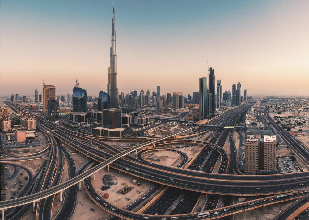 An aerial photograph of the Dubai skyline at sunset, featuring the iconic Burj Khalifa towering over other skyscrapers, with a complex web of intersecting highways and overpasses in the foreground, all under a gradient sky of orange and blue. The photo is displayed within a silver frame. Poster