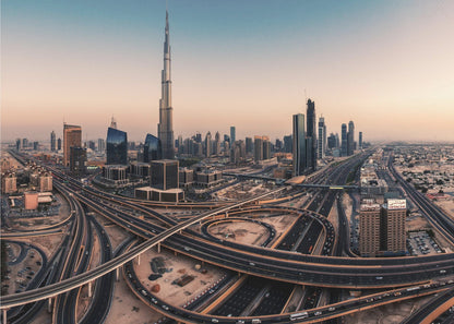 An aerial photograph of the Dubai skyline at sunset, featuring the iconic Burj Khalifa towering over other skyscrapers, with a complex web of intersecting highways and overpasses in the foreground, all under a gradient sky of orange and blue. The photo is displayed within a silver frame. Poster