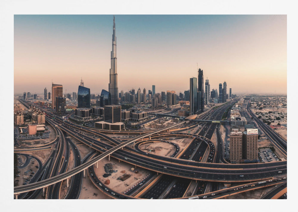 An aerial photograph of the Dubai skyline at sunset, featuring the iconic Burj Khalifa towering over other skyscrapers, with a complex web of intersecting highways and overpasses in the foreground, all under a gradient sky of orange and blue. The photo is displayed within a silver frame. Poster