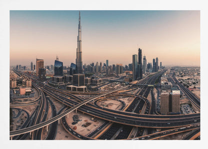 An aerial photograph of the Dubai skyline at sunset, featuring the iconic Burj Khalifa towering over other skyscrapers, with a complex web of intersecting highways and overpasses in the foreground, all under a gradient sky of orange and blue. The photo is displayed within a silver frame. Poster