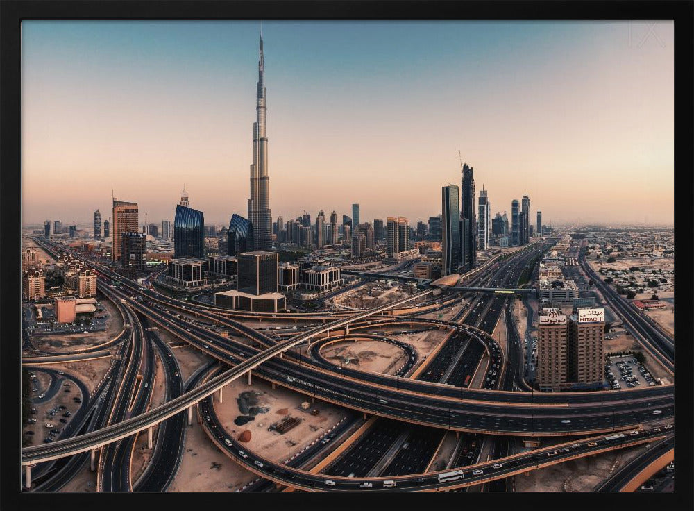 An aerial photograph of the Dubai skyline at sunset, featuring the iconic Burj Khalifa towering over other skyscrapers, with a complex web of intersecting highways and overpasses in the foreground, all under a gradient sky of orange and blue. The photo is displayed within a silver frame. Poster