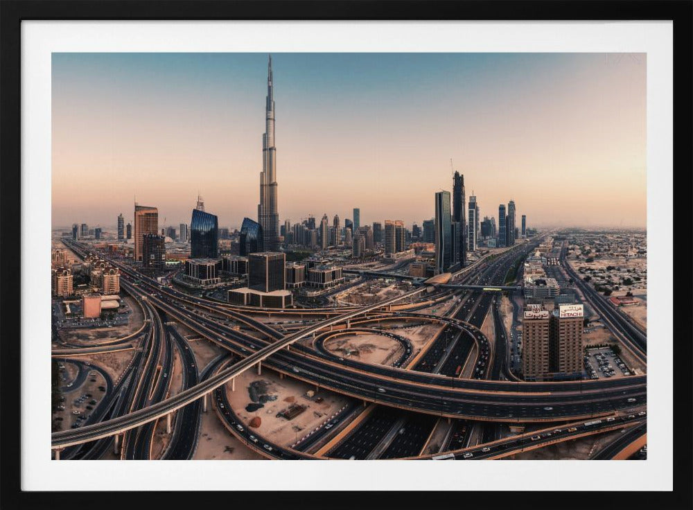 An aerial photograph of the Dubai skyline at sunset, featuring the iconic Burj Khalifa towering over other skyscrapers, with a complex web of intersecting highways and overpasses in the foreground, all under a gradient sky of orange and blue. The photo is displayed within a silver frame. Poster