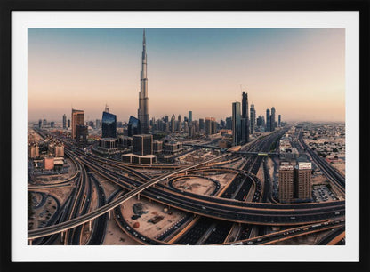 An aerial photograph of the Dubai skyline at sunset, featuring the iconic Burj Khalifa towering over other skyscrapers, with a complex web of intersecting highways and overpasses in the foreground, all under a gradient sky of orange and blue. The photo is displayed within a silver frame. Poster