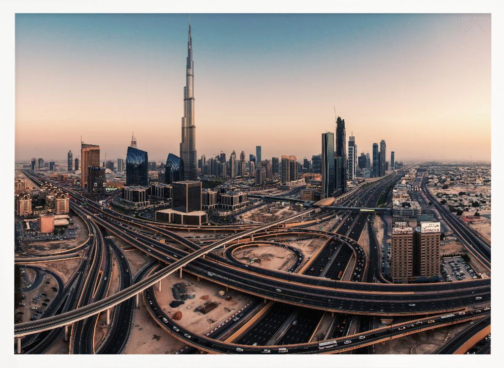 An aerial photograph of the Dubai skyline at sunset, featuring the iconic Burj Khalifa towering over other skyscrapers, with a complex web of intersecting highways and overpasses in the foreground, all under a gradient sky of orange and blue. The photo is displayed within a silver frame. Poster