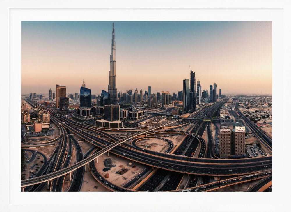 An aerial photograph of the Dubai skyline at sunset, featuring the iconic Burj Khalifa towering over other skyscrapers, with a complex web of intersecting highways and overpasses in the foreground, all under a gradient sky of orange and blue. The photo is displayed within a silver frame. Poster