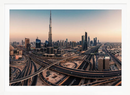 An aerial photograph of the Dubai skyline at sunset, featuring the iconic Burj Khalifa towering over other skyscrapers, with a complex web of intersecting highways and overpasses in the foreground, all under a gradient sky of orange and blue. The photo is displayed within a silver frame. Poster