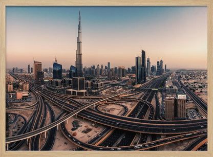 An aerial photograph of the Dubai skyline at sunset, featuring the iconic Burj Khalifa towering over other skyscrapers, with a complex web of intersecting highways and overpasses in the foreground, all under a gradient sky of orange and blue. The photo is displayed within a silver frame. Poster
