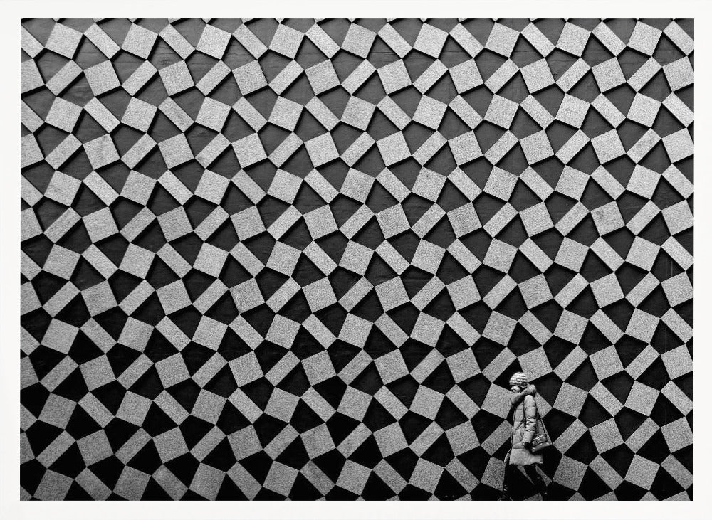 A black and white photograph of a lone person walking in front of a massive wall covered in a dizzying geometric pattern of tilted, textured gray quadrilaterals on a black background. The figure in the bottom right corner is dwarfed by the scale of the abstract architectural design. The image has a silver-gray frame. Artwork