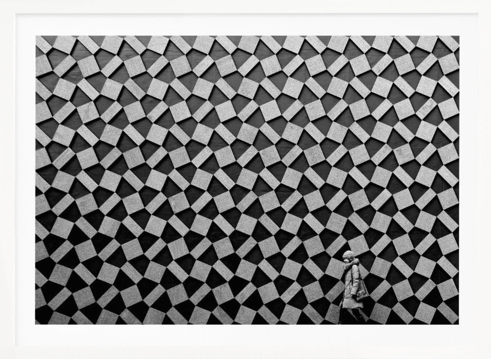 A black and white photograph of a lone person walking in front of a massive wall covered in a dizzying geometric pattern of tilted, textured gray quadrilaterals on a black background. The figure in the bottom right corner is dwarfed by the scale of the abstract architectural design. The image has a silver-gray frame. Artwork