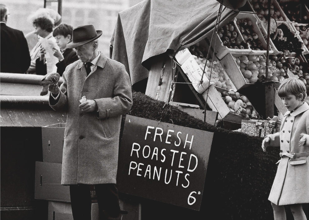 A vintage black and white photograph of an elderly man in a fedora and overcoat feeding a pigeon from his hand at an outdoor market. A sign for 'FRESH ROASTED PEANUTS' is visible next to him, and a young boy in a coat watches nearby. Wall Art