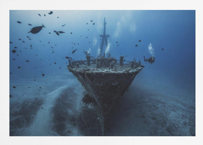 An underwater photograph of a large, sunken shipwreck resting on the sandy ocean floor. Two scuba divers are exploring the wreck, surrounded by schools of small fish in the deep blue water, with light filtering down from the surface. Print