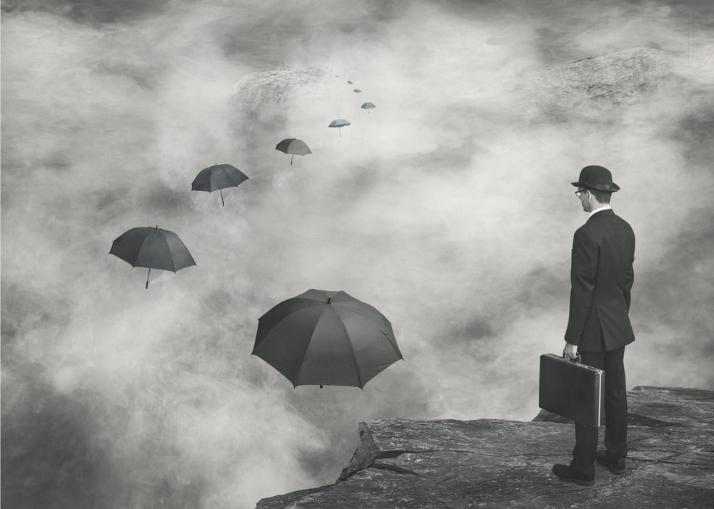 A surreal black and white photograph of a man in a suit and bowler hat holding a briefcase, standing on a cliff edge. He gazes into a foggy expanse where a path of floating black umbrellas leads into the misty distance. Decor