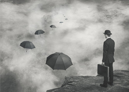 A surreal black and white photograph of a man in a suit and bowler hat holding a briefcase, standing on a cliff edge. He gazes into a foggy expanse where a path of floating black umbrellas leads into the misty distance. Decor