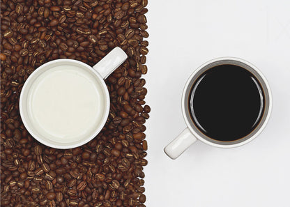 A top-down, split-screen photograph showing a contrast between coffee and milk. On the left, a white mug of milk sits on a bed of roasted coffee beans. On the right, a white mug of black coffee rests on a clean white background, creating a stark visual divide. Artwork