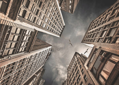 A dramatic, low-angle photograph looking up at the sky between several tall, sepia-toned skyscrapers that converge towards the center. Two birds are seen flying in the moody, dark grey sky. The image has a vintage feel and is presented in a silver frame. Wall Art