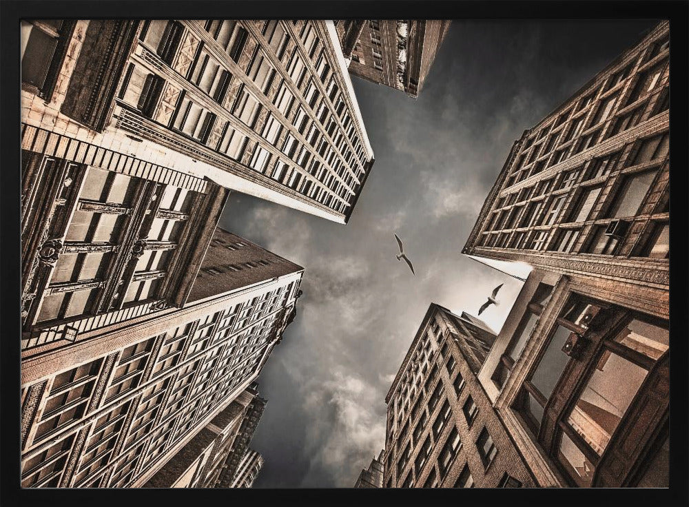 A dramatic, low-angle photograph looking up at the sky between several tall, sepia-toned skyscrapers that converge towards the center. Two birds are seen flying in the moody, dark grey sky. The image has a vintage feel and is presented in a silver frame. Wall Art