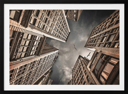 A dramatic, low-angle photograph looking up at the sky between several tall, sepia-toned skyscrapers that converge towards the center. Two birds are seen flying in the moody, dark grey sky. The image has a vintage feel and is presented in a silver frame. Wall Art