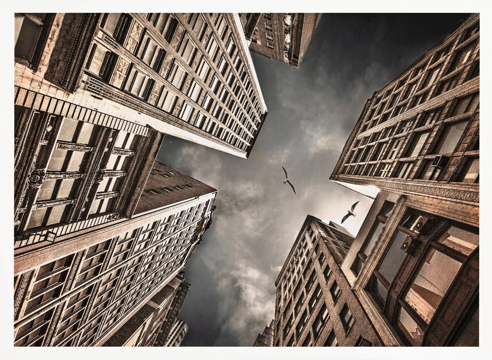 A dramatic, low-angle photograph looking up at the sky between several tall, sepia-toned skyscrapers that converge towards the center. Two birds are seen flying in the moody, dark grey sky. The image has a vintage feel and is presented in a silver frame. Wall Art