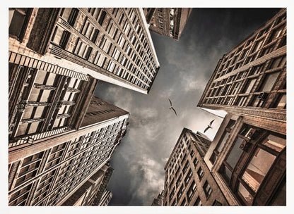 A dramatic, low-angle photograph looking up at the sky between several tall, sepia-toned skyscrapers that converge towards the center. Two birds are seen flying in the moody, dark grey sky. The image has a vintage feel and is presented in a silver frame. Wall Art