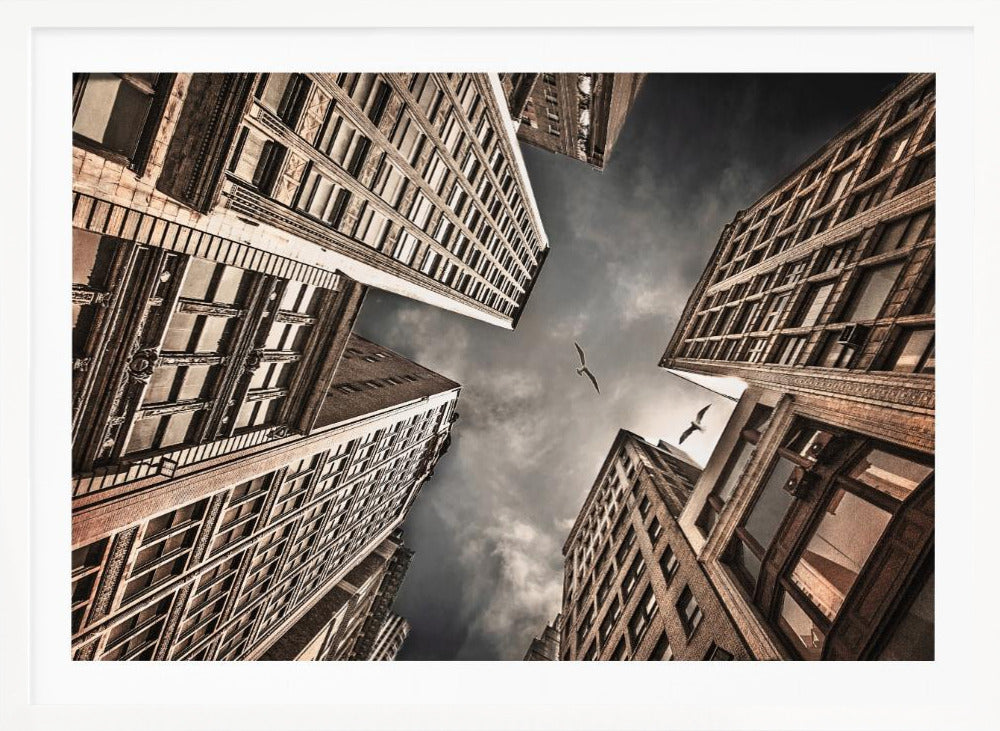 A dramatic, low-angle photograph looking up at the sky between several tall, sepia-toned skyscrapers that converge towards the center. Two birds are seen flying in the moody, dark grey sky. The image has a vintage feel and is presented in a silver frame. Wall Art