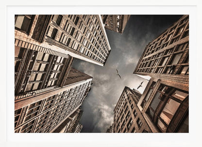 A dramatic, low-angle photograph looking up at the sky between several tall, sepia-toned skyscrapers that converge towards the center. Two birds are seen flying in the moody, dark grey sky. The image has a vintage feel and is presented in a silver frame. Wall Art