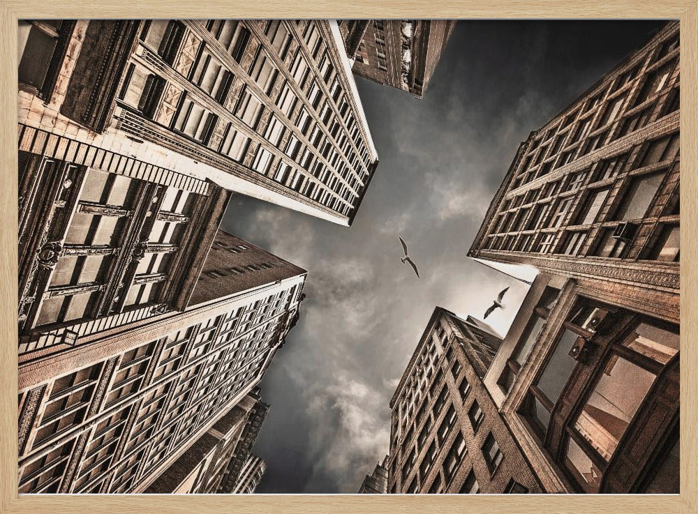 A dramatic, low-angle photograph looking up at the sky between several tall, sepia-toned skyscrapers that converge towards the center. Two birds are seen flying in the moody, dark grey sky. The image has a vintage feel and is presented in a silver frame. Wall Art
