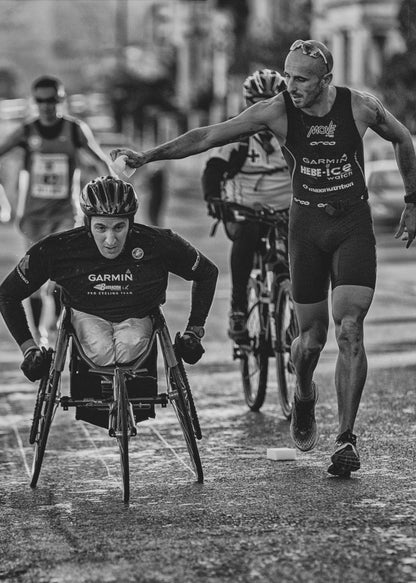 A dynamic black and white action photograph of a para-athlete in a racing wheelchair during a race. A running athlete beside him reaches out to hand him a sponge, showcasing a moment of camaraderie and support. Other competitors are blurred in the background on the wet asphalt. Print