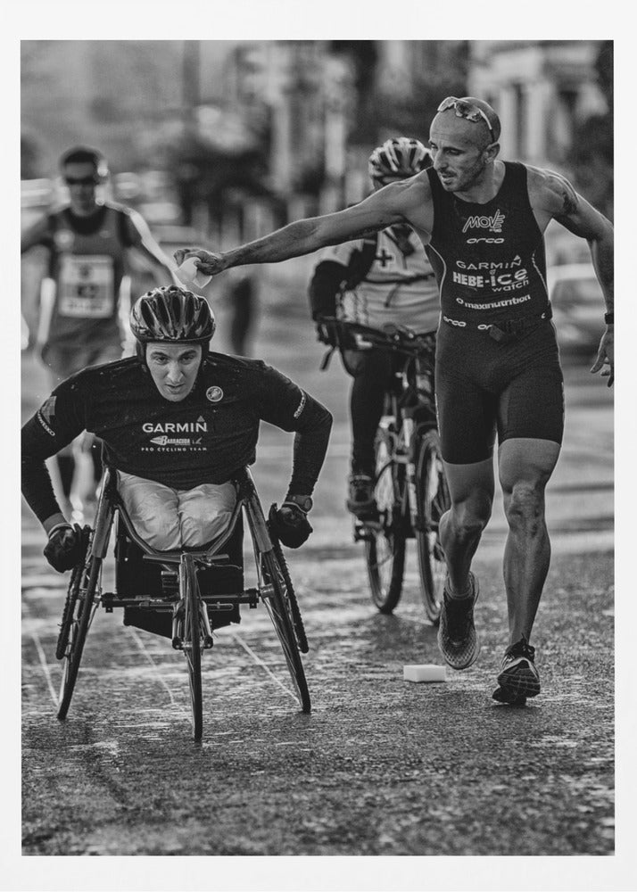 A dynamic black and white action photograph of a para-athlete in a racing wheelchair during a race. A running athlete beside him reaches out to hand him a sponge, showcasing a moment of camaraderie and support. Other competitors are blurred in the background on the wet asphalt. Print