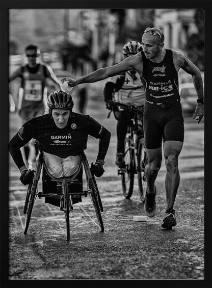 A dynamic black and white action photograph of a para-athlete in a racing wheelchair during a race. A running athlete beside him reaches out to hand him a sponge, showcasing a moment of camaraderie and support. Other competitors are blurred in the background on the wet asphalt. Print