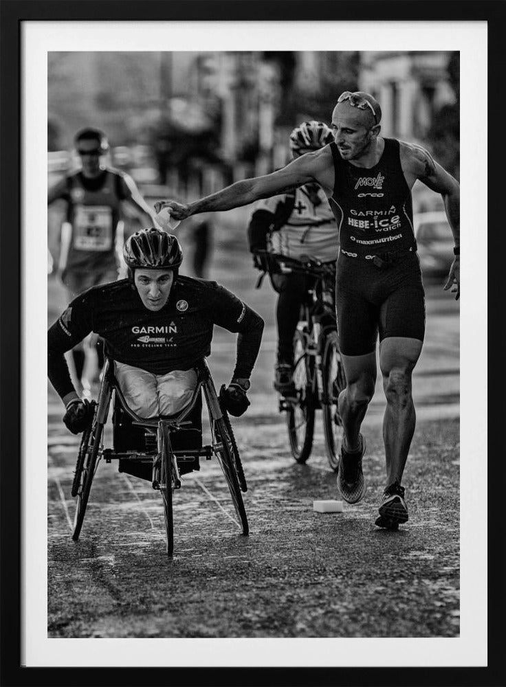 A dynamic black and white action photograph of a para-athlete in a racing wheelchair during a race. A running athlete beside him reaches out to hand him a sponge, showcasing a moment of camaraderie and support. Other competitors are blurred in the background on the wet asphalt. Print