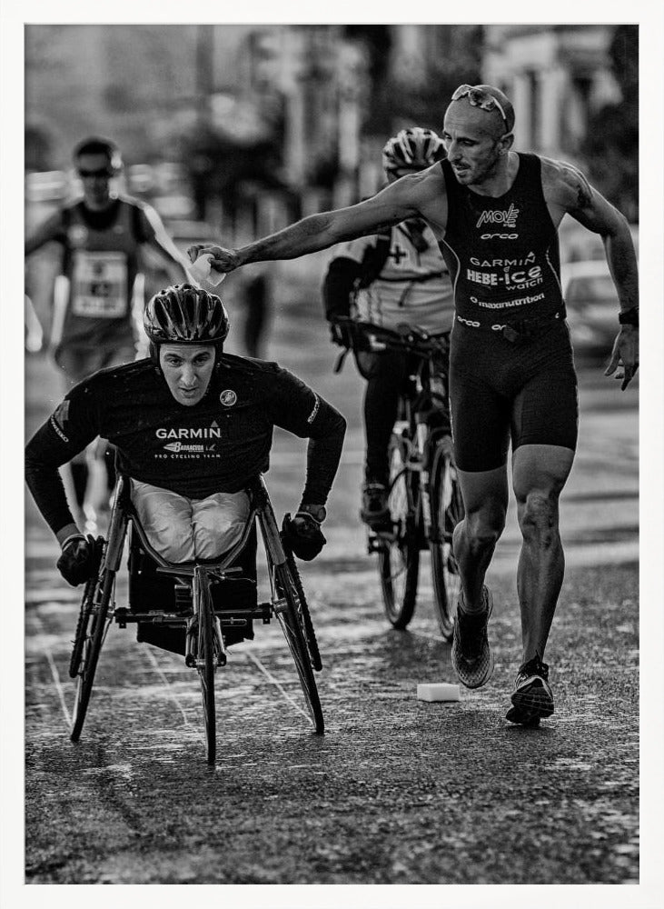A dynamic black and white action photograph of a para-athlete in a racing wheelchair during a race. A running athlete beside him reaches out to hand him a sponge, showcasing a moment of camaraderie and support. Other competitors are blurred in the background on the wet asphalt. Print