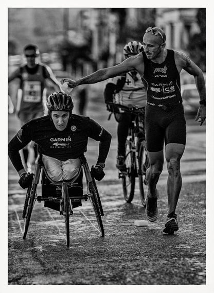A dynamic black and white action photograph of a para-athlete in a racing wheelchair during a race. A running athlete beside him reaches out to hand him a sponge, showcasing a moment of camaraderie and support. Other competitors are blurred in the background on the wet asphalt. Print