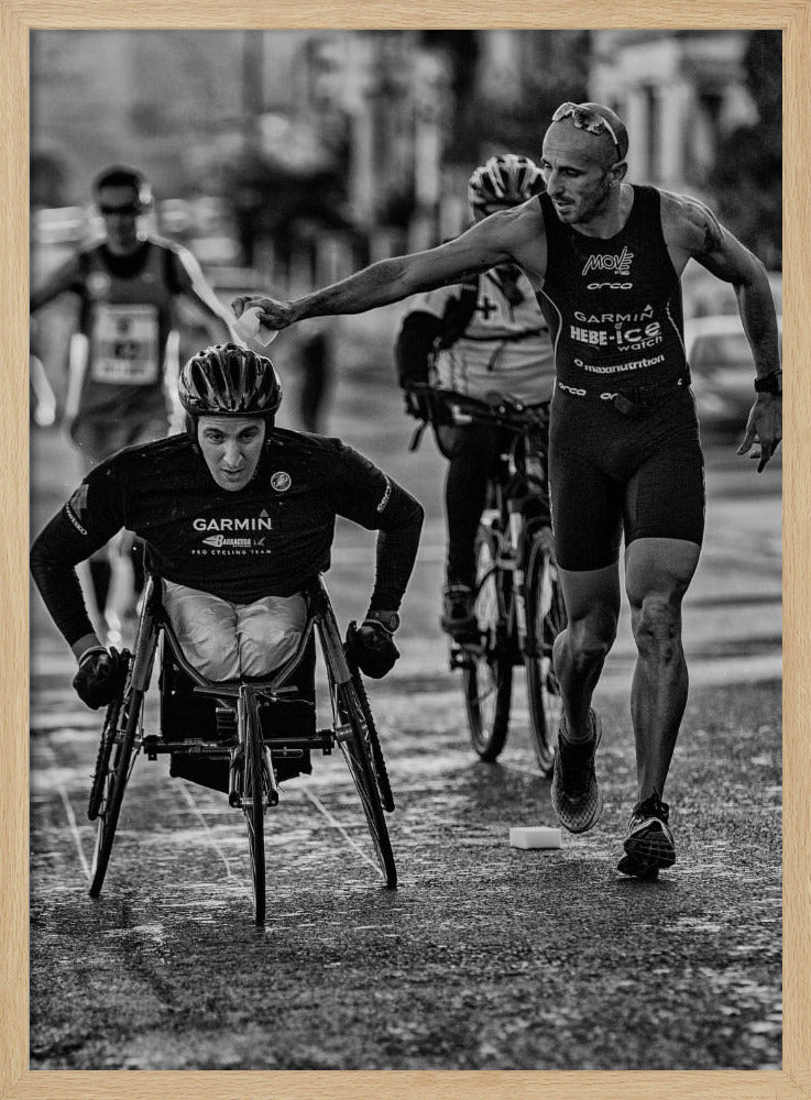 A dynamic black and white action photograph of a para-athlete in a racing wheelchair during a race. A running athlete beside him reaches out to hand him a sponge, showcasing a moment of camaraderie and support. Other competitors are blurred in the background on the wet asphalt. Print