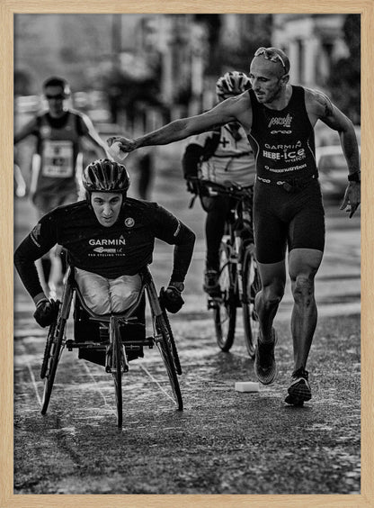A dynamic black and white action photograph of a para-athlete in a racing wheelchair during a race. A running athlete beside him reaches out to hand him a sponge, showcasing a moment of camaraderie and support. Other competitors are blurred in the background on the wet asphalt. Print