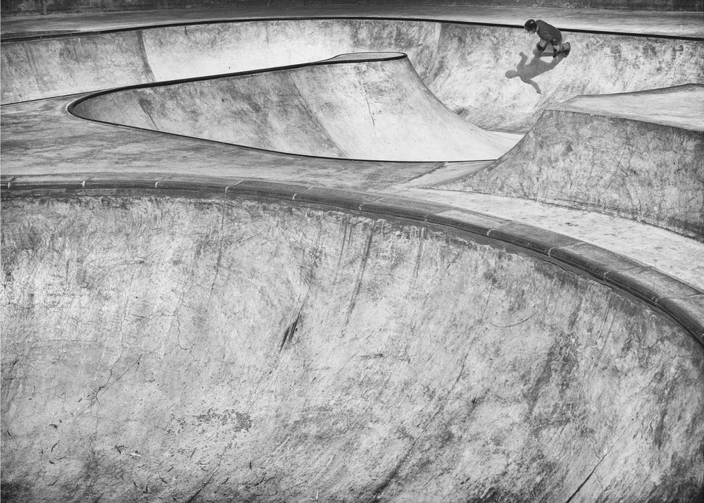 A high-angle, black and white photograph of a sprawling concrete skatepark with smooth, curving ramps. A single skateboarder is visible in the upper right, casting a distinct shadow on the ramp as they ride. The image is enclosed in a silver frame. Decor