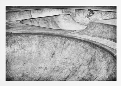 A high-angle, black and white photograph of a sprawling concrete skatepark with smooth, curving ramps. A single skateboarder is visible in the upper right, casting a distinct shadow on the ramp as they ride. The image is enclosed in a silver frame. Decor