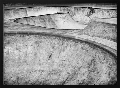 A high-angle, black and white photograph of a sprawling concrete skatepark with smooth, curving ramps. A single skateboarder is visible in the upper right, casting a distinct shadow on the ramp as they ride. The image is enclosed in a silver frame. Decor