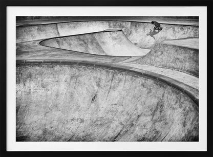 A high-angle, black and white photograph of a sprawling concrete skatepark with smooth, curving ramps. A single skateboarder is visible in the upper right, casting a distinct shadow on the ramp as they ride. The image is enclosed in a silver frame. Decor