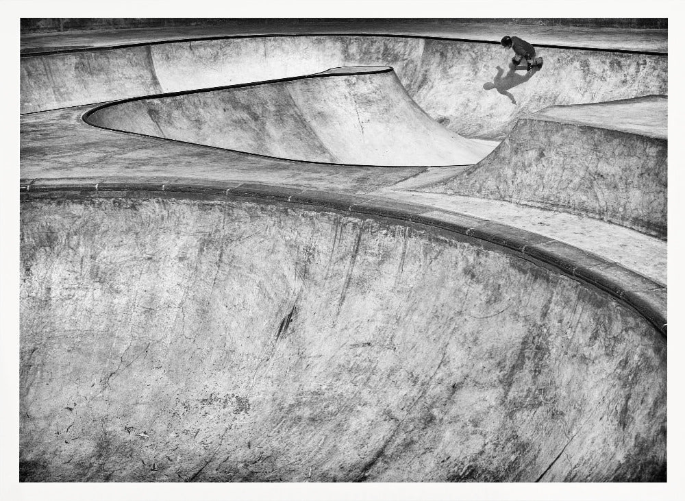 A high-angle, black and white photograph of a sprawling concrete skatepark with smooth, curving ramps. A single skateboarder is visible in the upper right, casting a distinct shadow on the ramp as they ride. The image is enclosed in a silver frame. Decor
