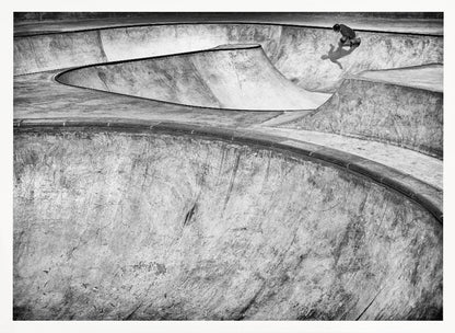 A high-angle, black and white photograph of a sprawling concrete skatepark with smooth, curving ramps. A single skateboarder is visible in the upper right, casting a distinct shadow on the ramp as they ride. The image is enclosed in a silver frame. Decor