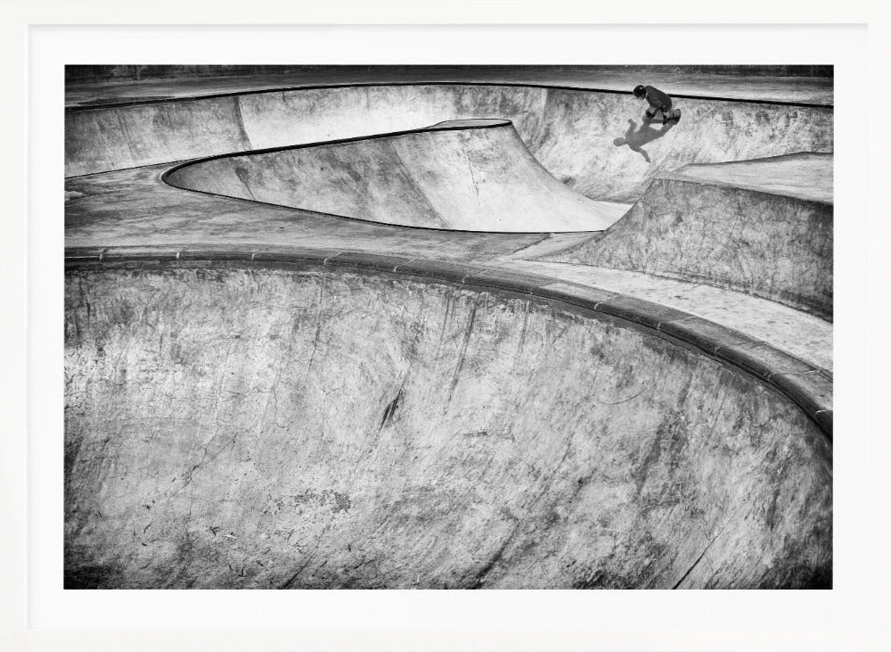 A high-angle, black and white photograph of a sprawling concrete skatepark with smooth, curving ramps. A single skateboarder is visible in the upper right, casting a distinct shadow on the ramp as they ride. The image is enclosed in a silver frame. Decor