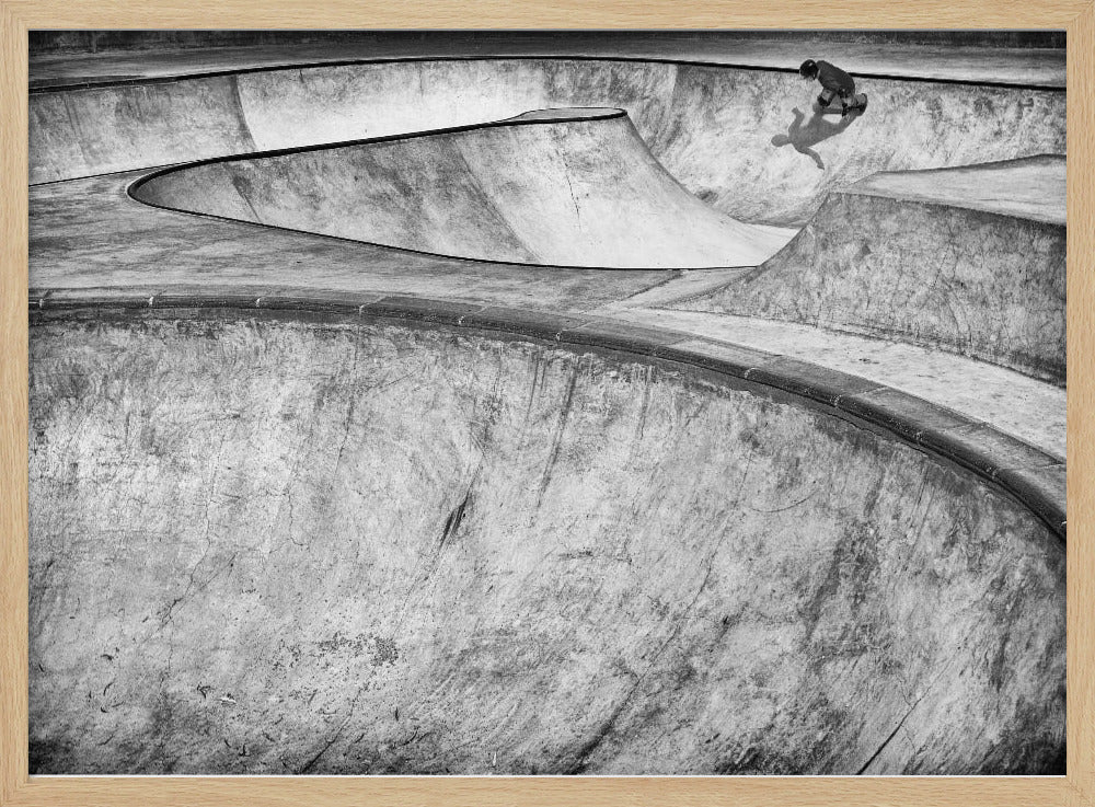 A high-angle, black and white photograph of a sprawling concrete skatepark with smooth, curving ramps. A single skateboarder is visible in the upper right, casting a distinct shadow on the ramp as they ride. The image is enclosed in a silver frame. Decor